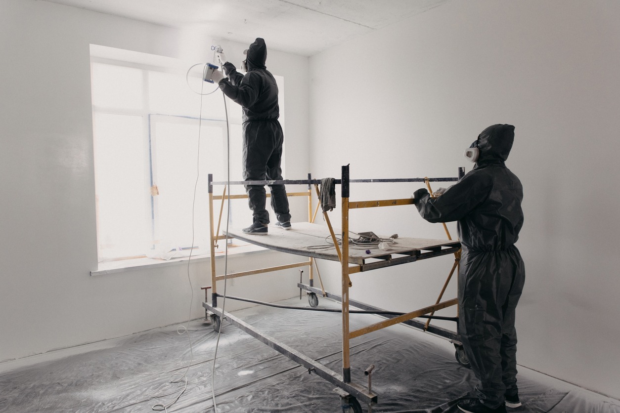 Male painters in protective suit is painting wall on stepladder with spray gun