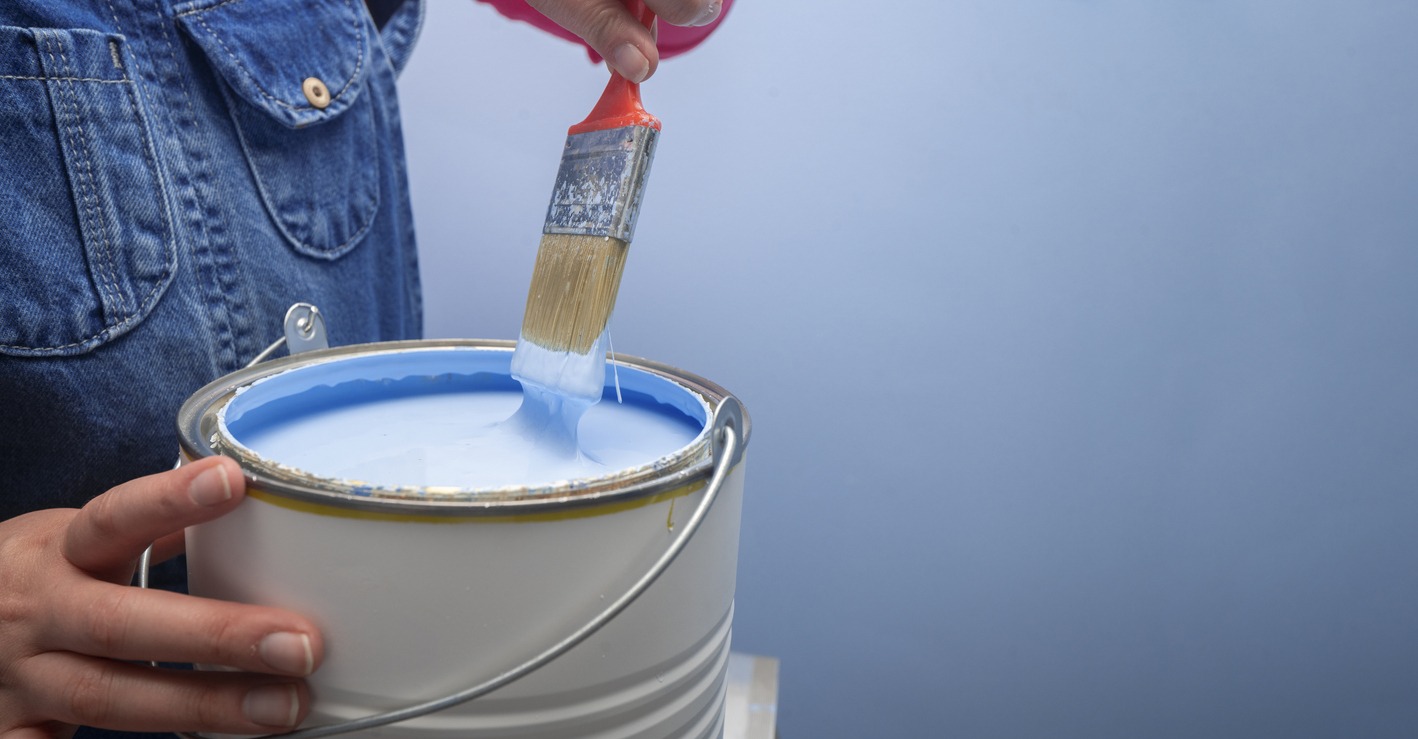 woman wearing overalls taking out the brush from the jar of blue paint next to a blue wall