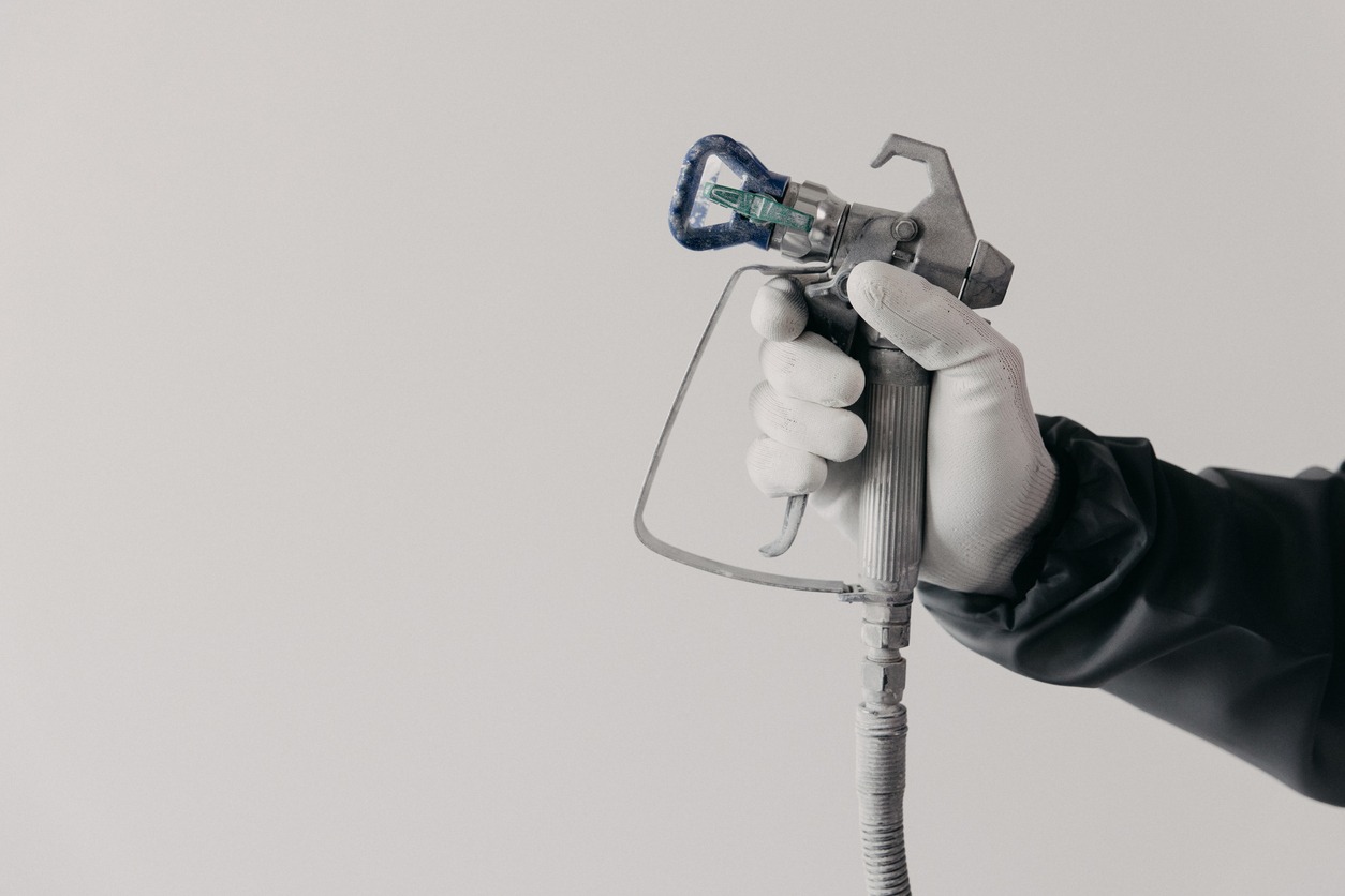 A painter's arm and hand in safety coveralls and gloves holding an Industrial size airless spray gun used for industrial painting and coating. Shot on white background.