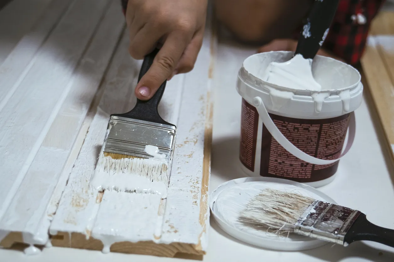 The boy holds a white paintbrush in his hand and paints a wooden surface.