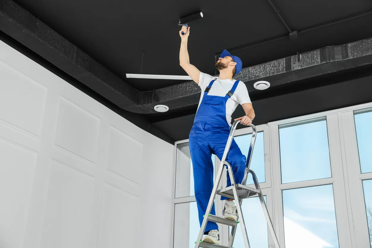 Worker in uniform painting ceiling with roller indoors, low angle view