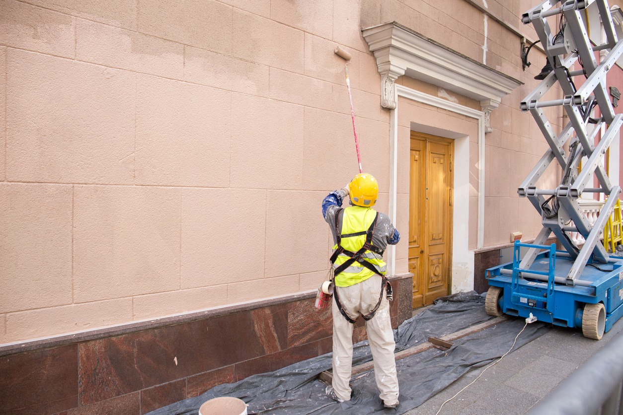 A rear view of a painter in a helmet painting a wall, with a paint roller and a bucket