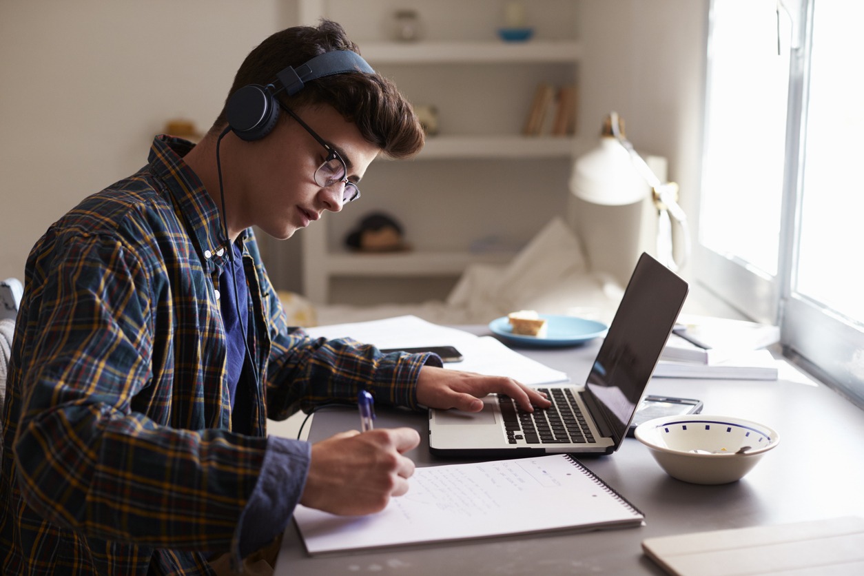 Teenage boy wearing headphones works at desk in his bedroom