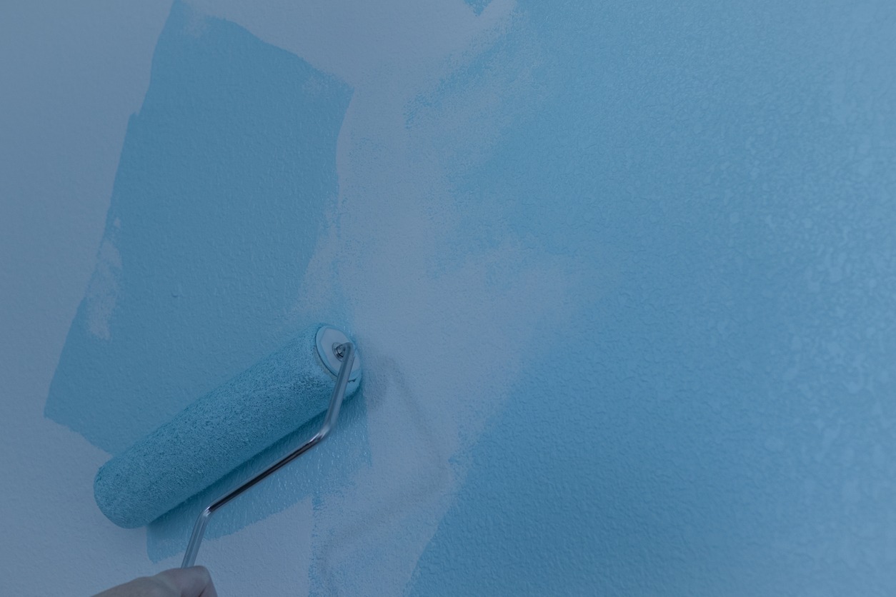 A hand rolling blue paint onto a white wall with a paint roller on an interior wall.