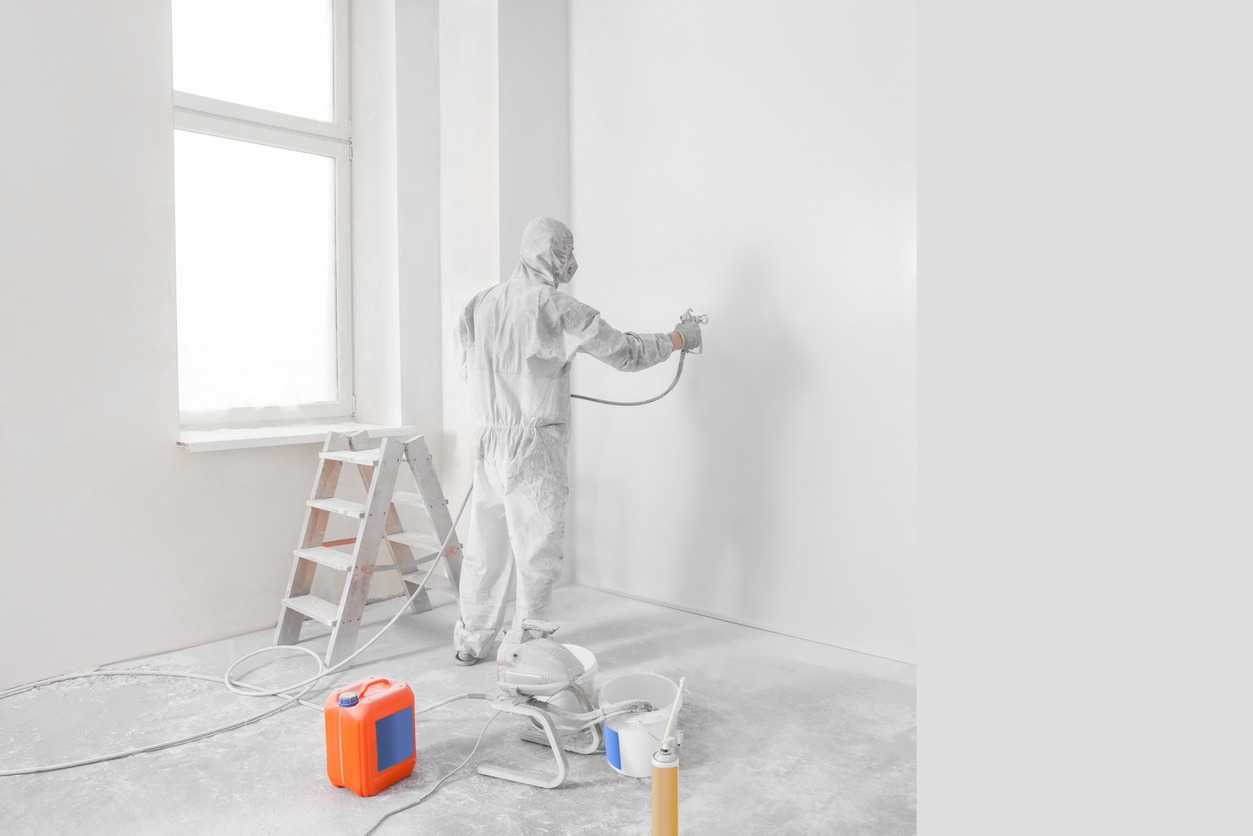 Decorator in uniform painting wall with sprayer indoors